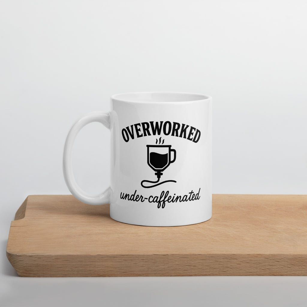 White mug with black text that says, “Overworked under-caffeinated,” featuring an illustration of a coffee cup connected to an IV line.