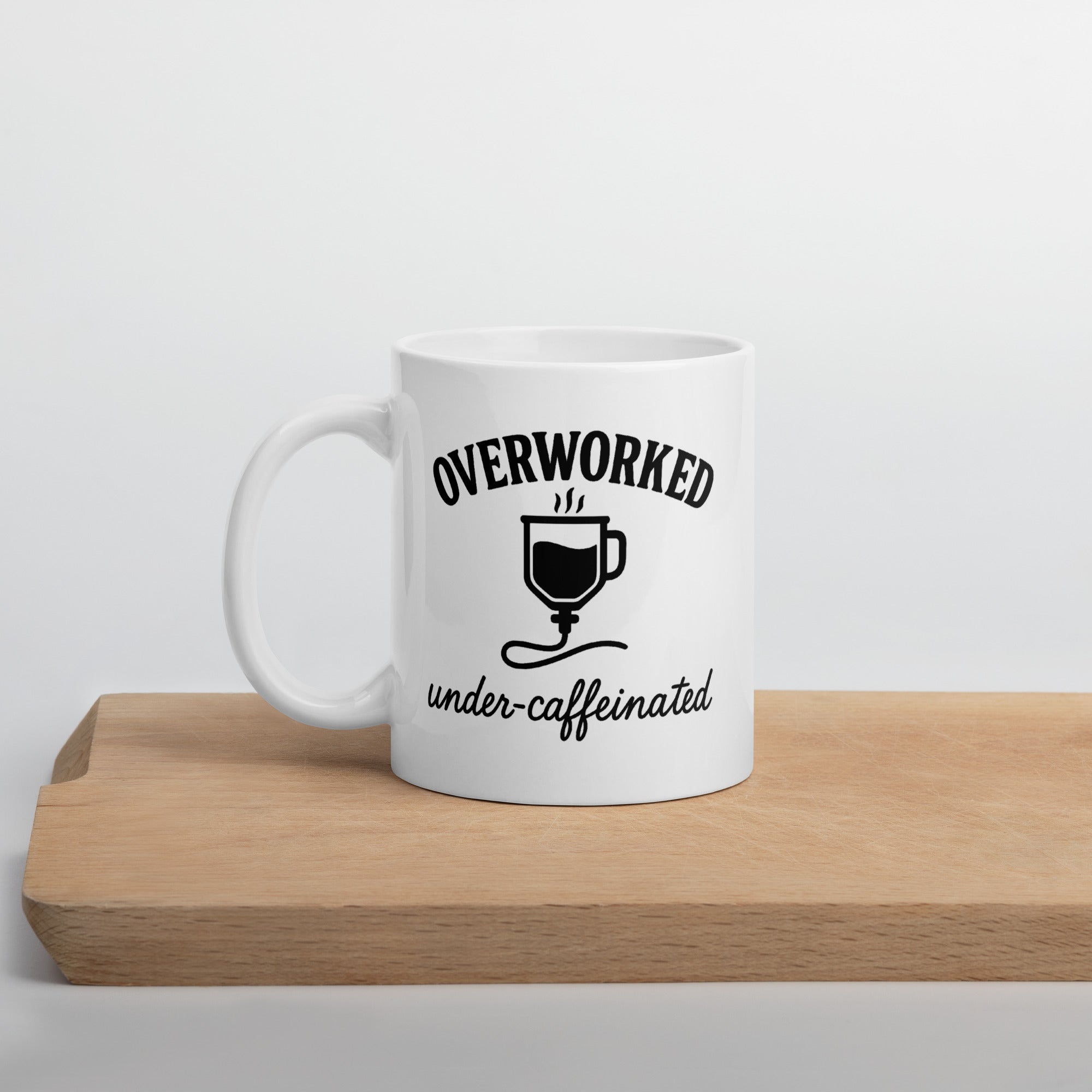 White mug with black text that says, “Overworked under-caffeinated,” featuring an illustration of a coffee cup connected to an IV line.