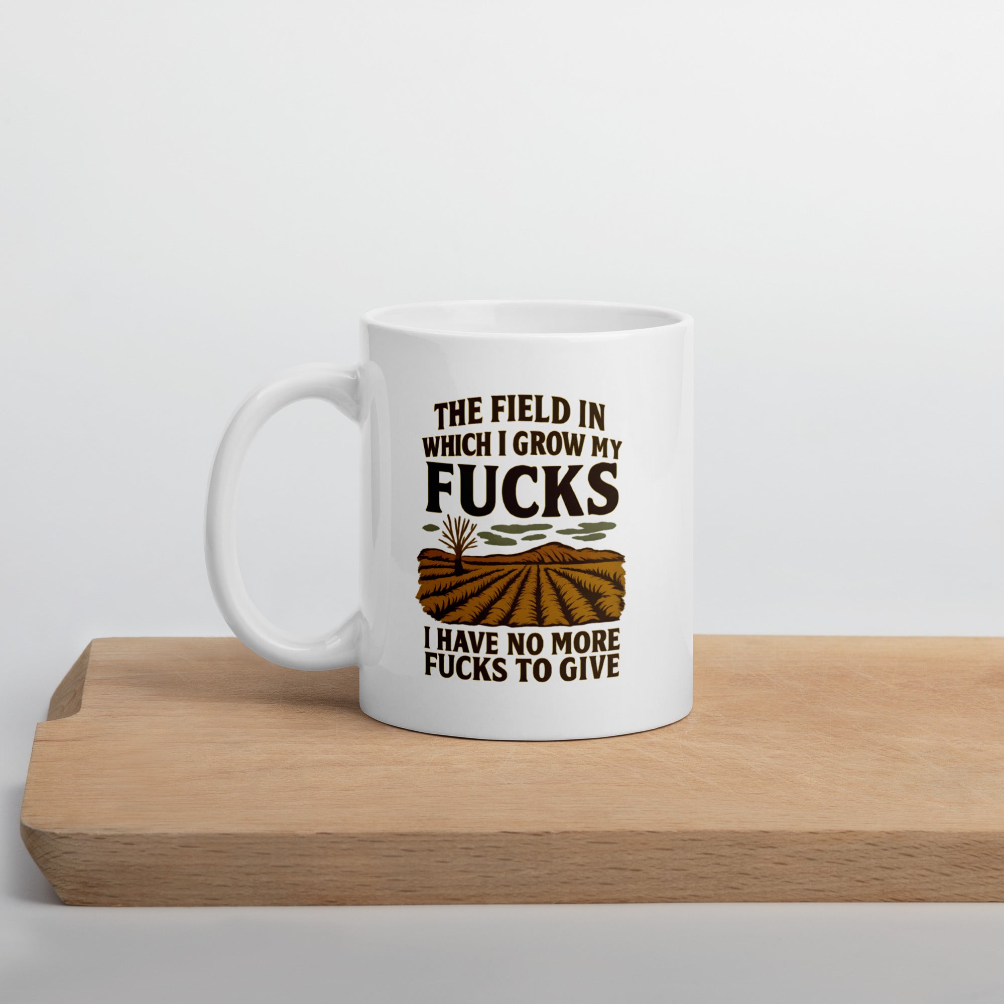 White coffee mug with a humorous design that reads, “The field in which I grow my fucks. I have no more fucks to give,” above and below an illustration of a barren field.